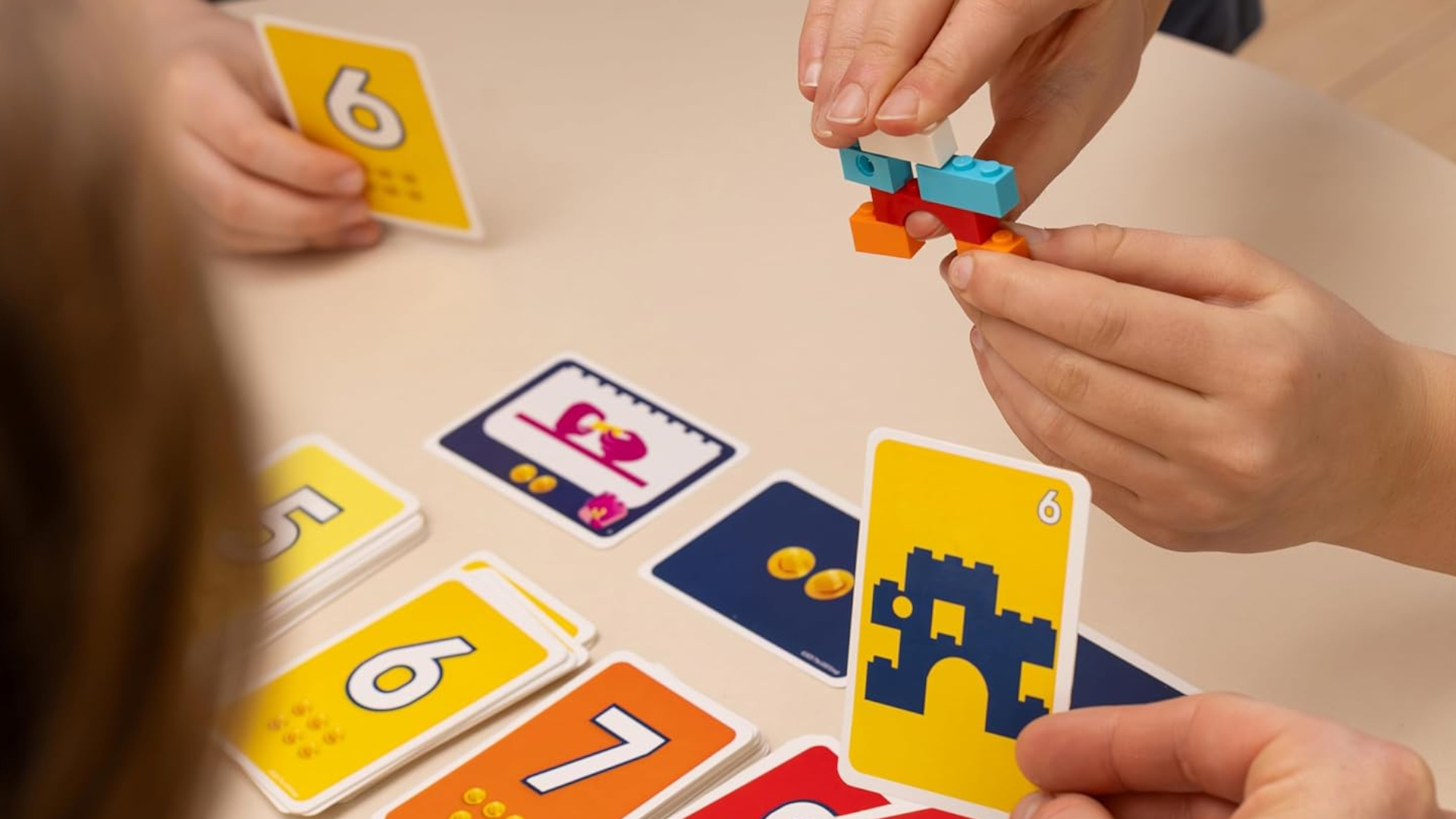 Hands piece together Lego bricks, while other hands hold onto cards at a table