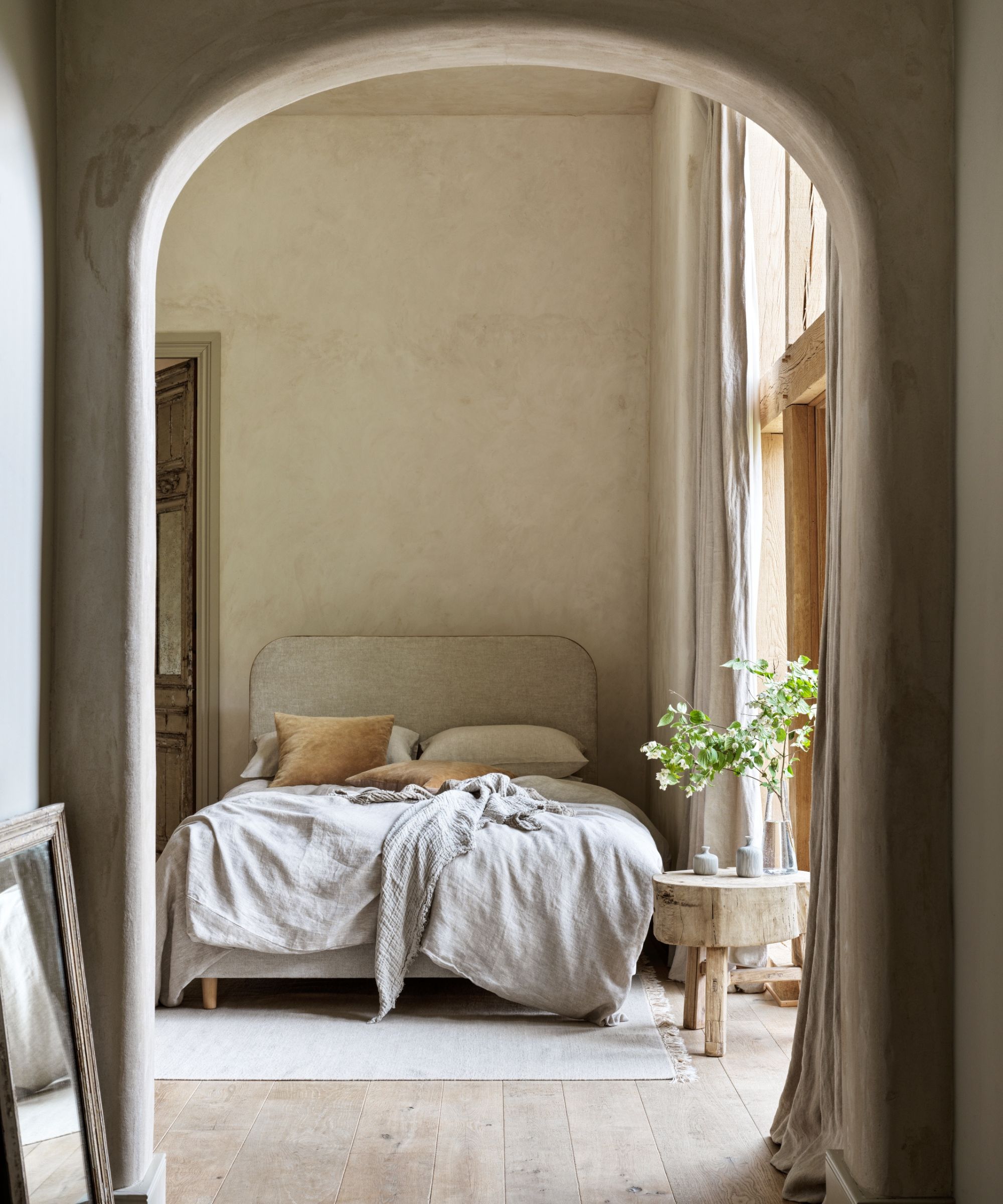 Viewed through a soft, rounded archway, is a bedroom with warm neutral tones. A low bed with textured beige bedding and tan cushions rests against a simple wall. Beside it, a small rustic wooden table holds a vase of leafy branches near a tall window draped in linen curtains.