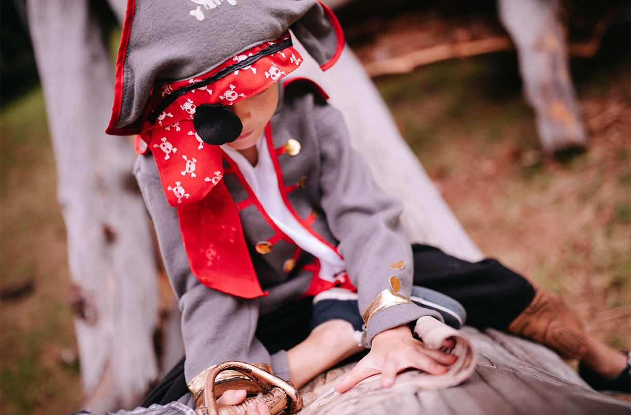 Little Boy dressing up as pirate sitting on log in park reading treasure map and holding saber in other hand