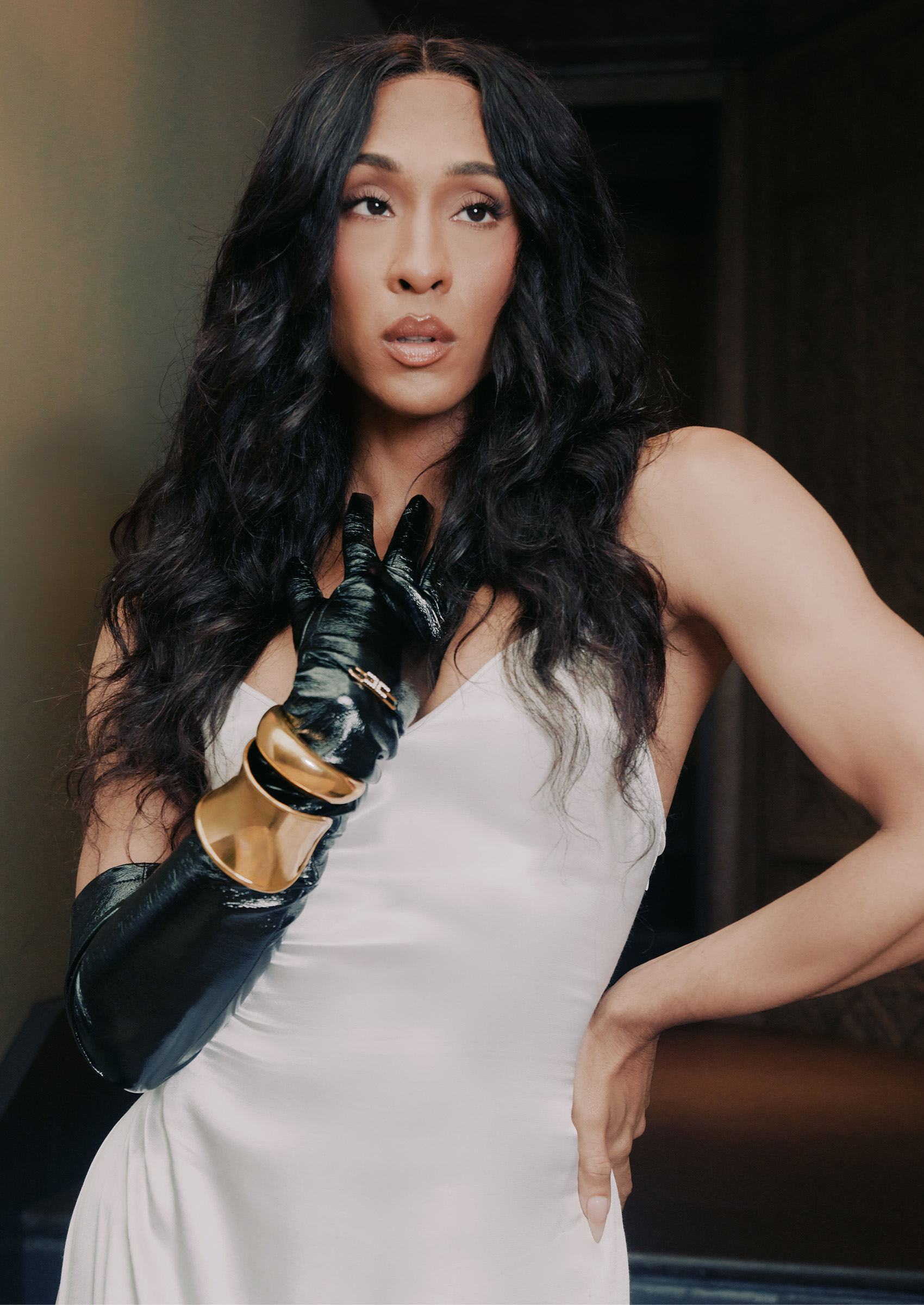 michaela jae rodriguez wears a white satin dress and a black glove with bracelets on one hand that she holds to her chest