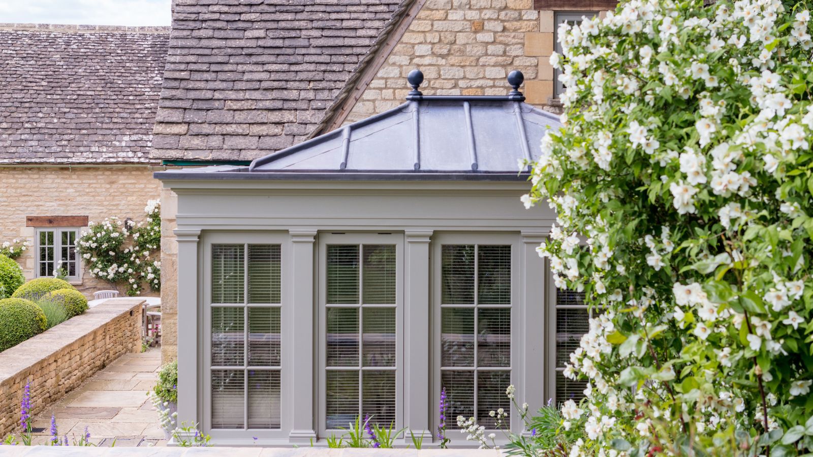 A neat grey painted orangery with a lead hip roof, black ball finials and full-height sash windows with venetian blinds.