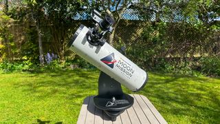 The Celestron Moon Mission 100mm Tabletop Dobsonian on a grey table with grass and trees in the background.