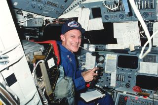 a smiling man in a blue flight suit sits in the space shuttle's cockpit, surrounded by buttons, levers and other controls.