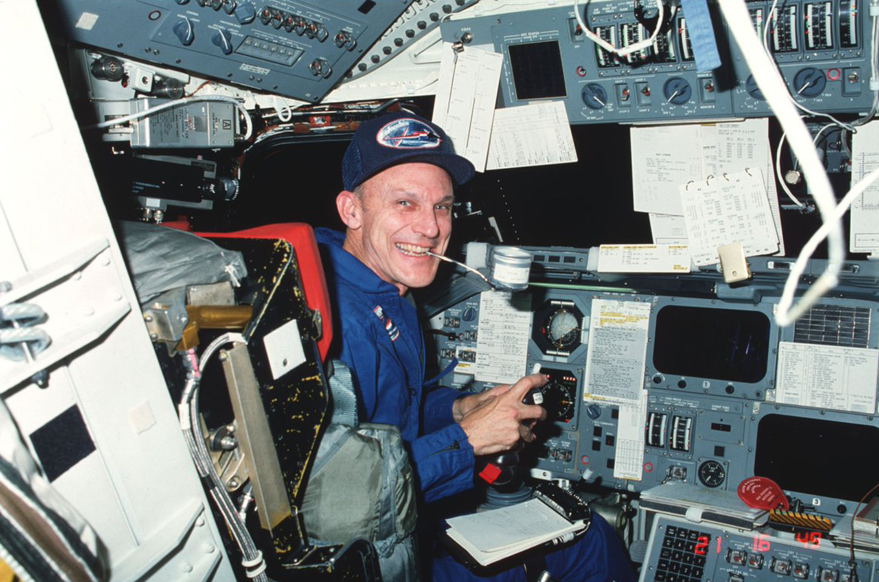a smiling man in a blue flight suit sits in the space shuttle's cockpit, surrounded by buttons, levers and other controls.