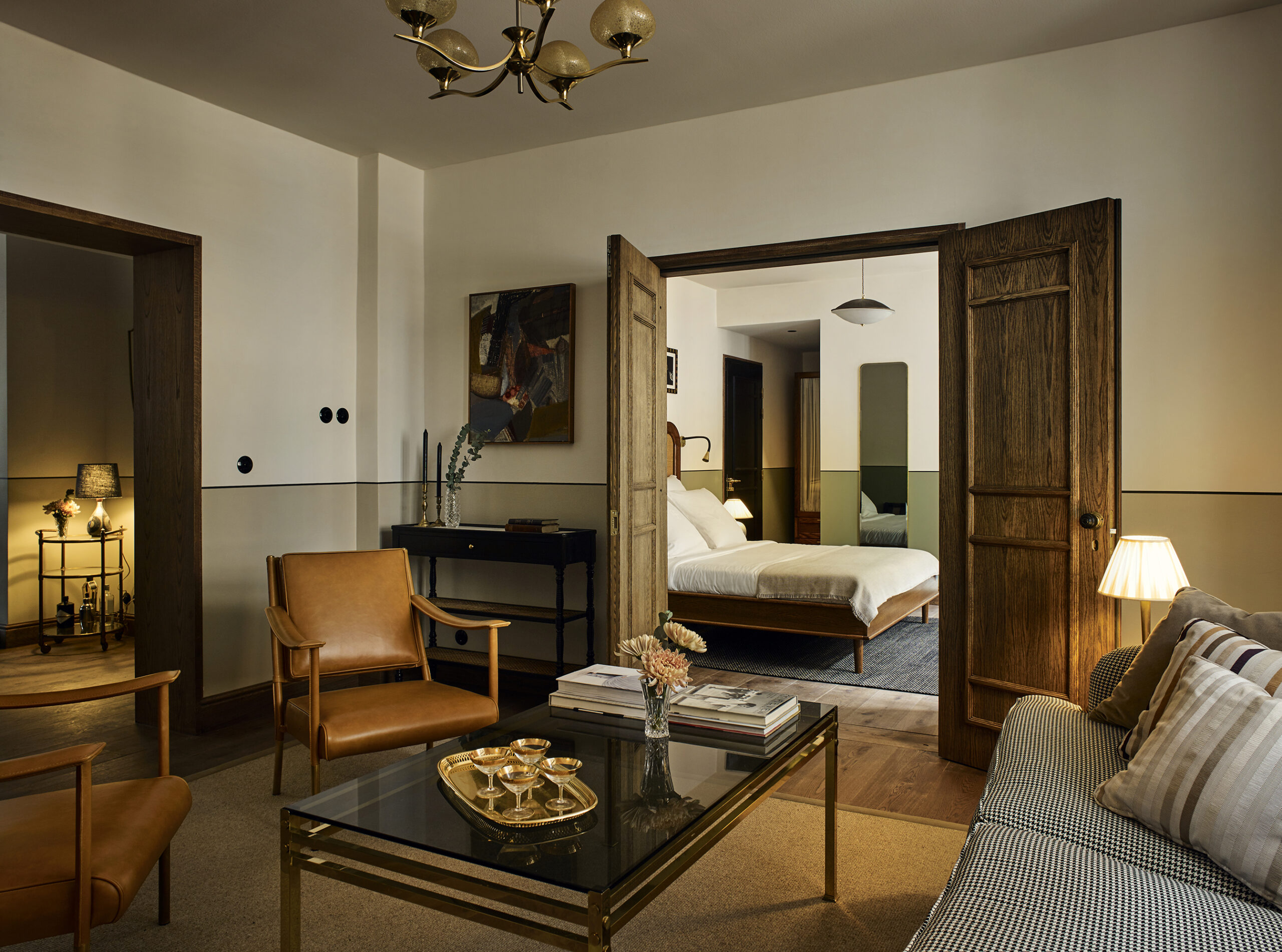 A golden-lit sprawling hotel suite is furnished with mid-century modern furniture in pale brown leather mixed with Art Deco accents like brass chandeliers, table lamps, and serveware.