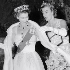 A black and white photo of Queen Elizabeth II wearing an evening gown and tiara getting a fur stole adjusted by Pamela Mountbatten