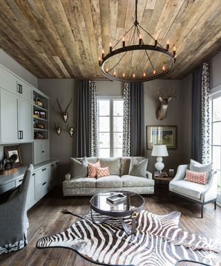 Dark wooden ceiling, rounded black metal chandelier, gray painted walls, zebra print rug, gray sofa and armchair, built in gray storage cabinet, blue patterned curtains