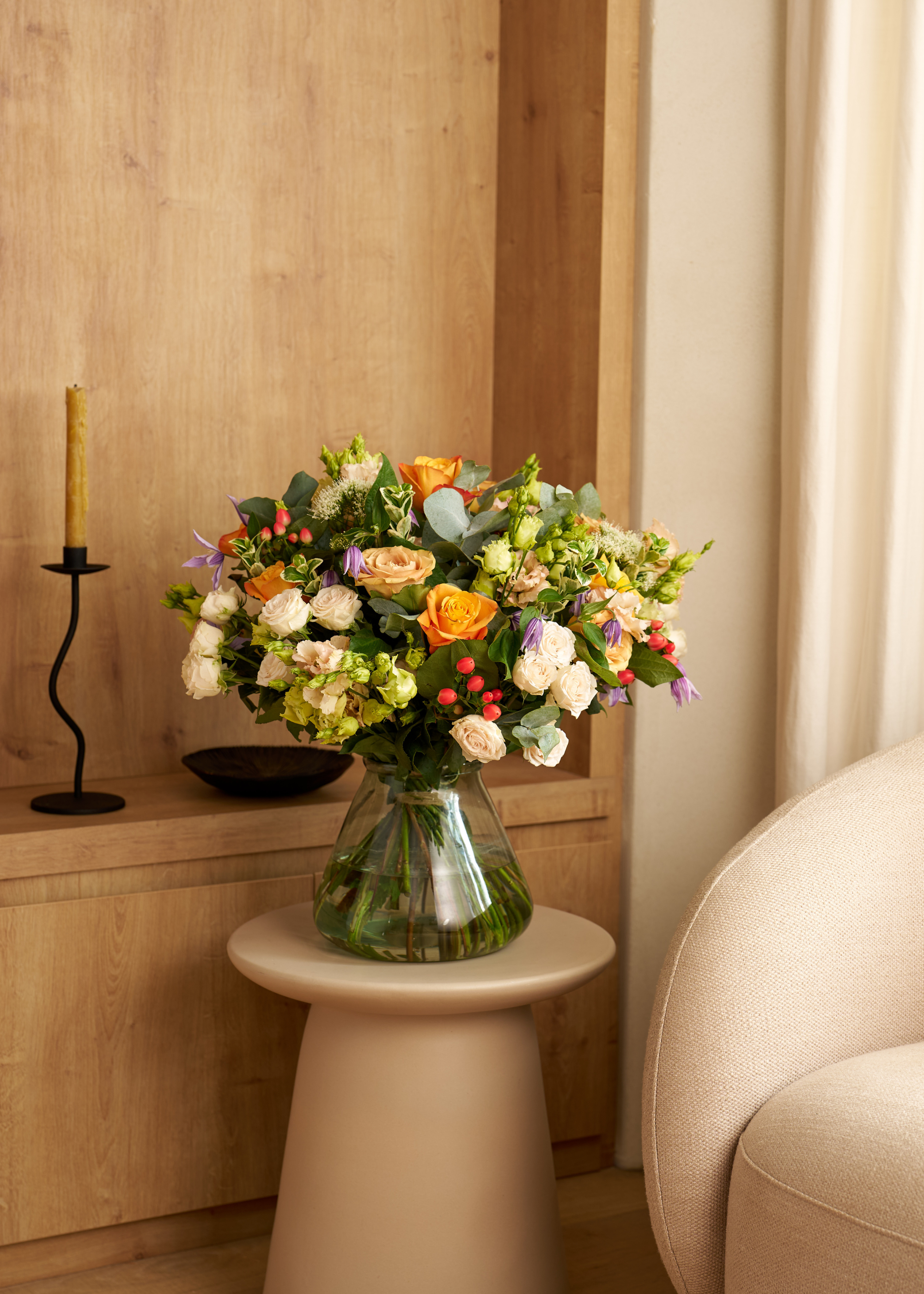 A living room corner with a cream armchair, a side table with a glass vase of white and peach roses with sweet peas beside a wood storage nook with a ceramic bowl and a wavy metal candlestick