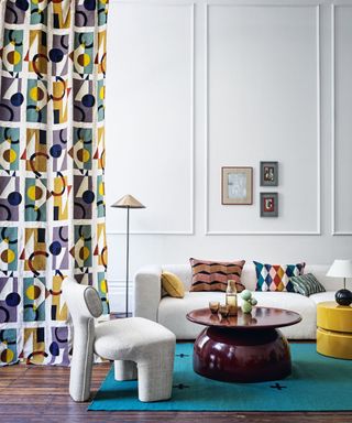 Modern living room with white painted walls and panels, blue and yellow geometric patterned curtain, dark wood flooring, blue rug, off-white chunky sofa and armchair, yellow ottoman, table lamp and floor lamp with cone shade, dark brown gloss rounded coffee table