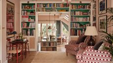 a large soft pink victorian 'middle room' with white and green bookcases built around a doorway looking into a large bright kitchen