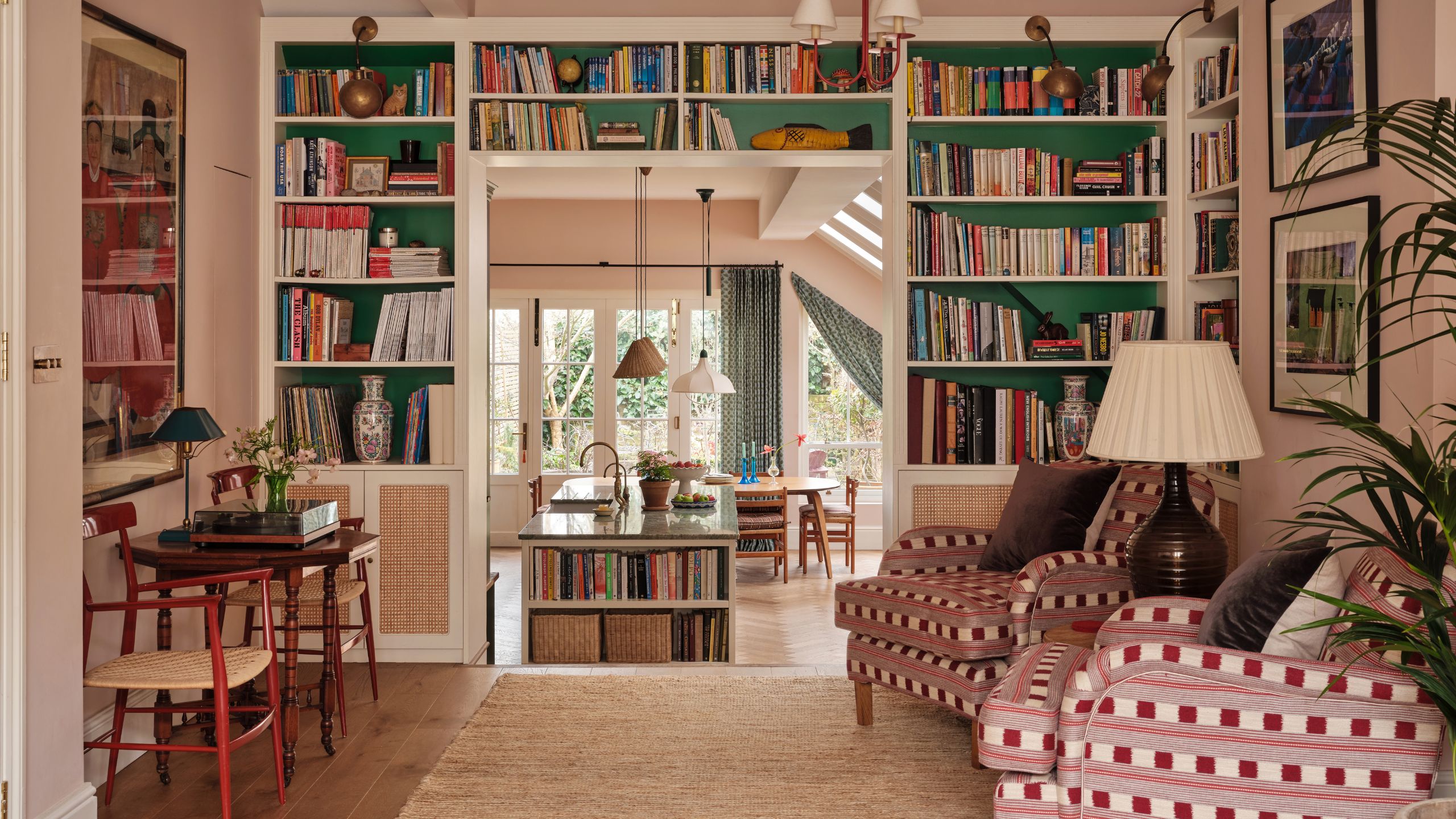 a large soft pink victorian 'middle room' with white and green bookcases built around a doorway looking into a large bright kitchen