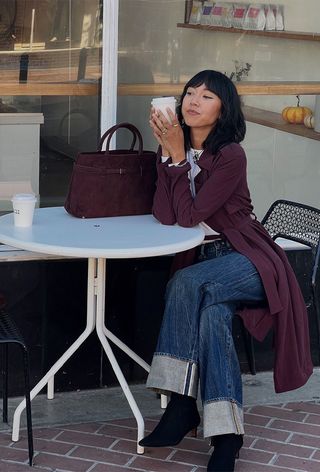 best suede boots shown on a photo of a woman sitting outside at a cafe wearing a red trench coat over a white t-shirt with wide-leg cuffed jeans, black suede ankle boots, and a red suede handbag