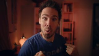 A man in a blue shirt with wide eyes and an excited expression points to himself in a warmly lit, dramatic indoor setting.