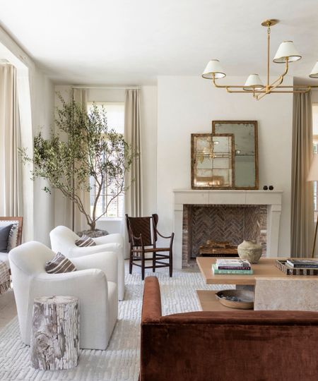 Transitional living room with soft white walls, beige curtains, a stone fireplace with antique mirrors, a light gray rug with two white armchairs, a wooden coffee table, and a rust-colored velvet sofa