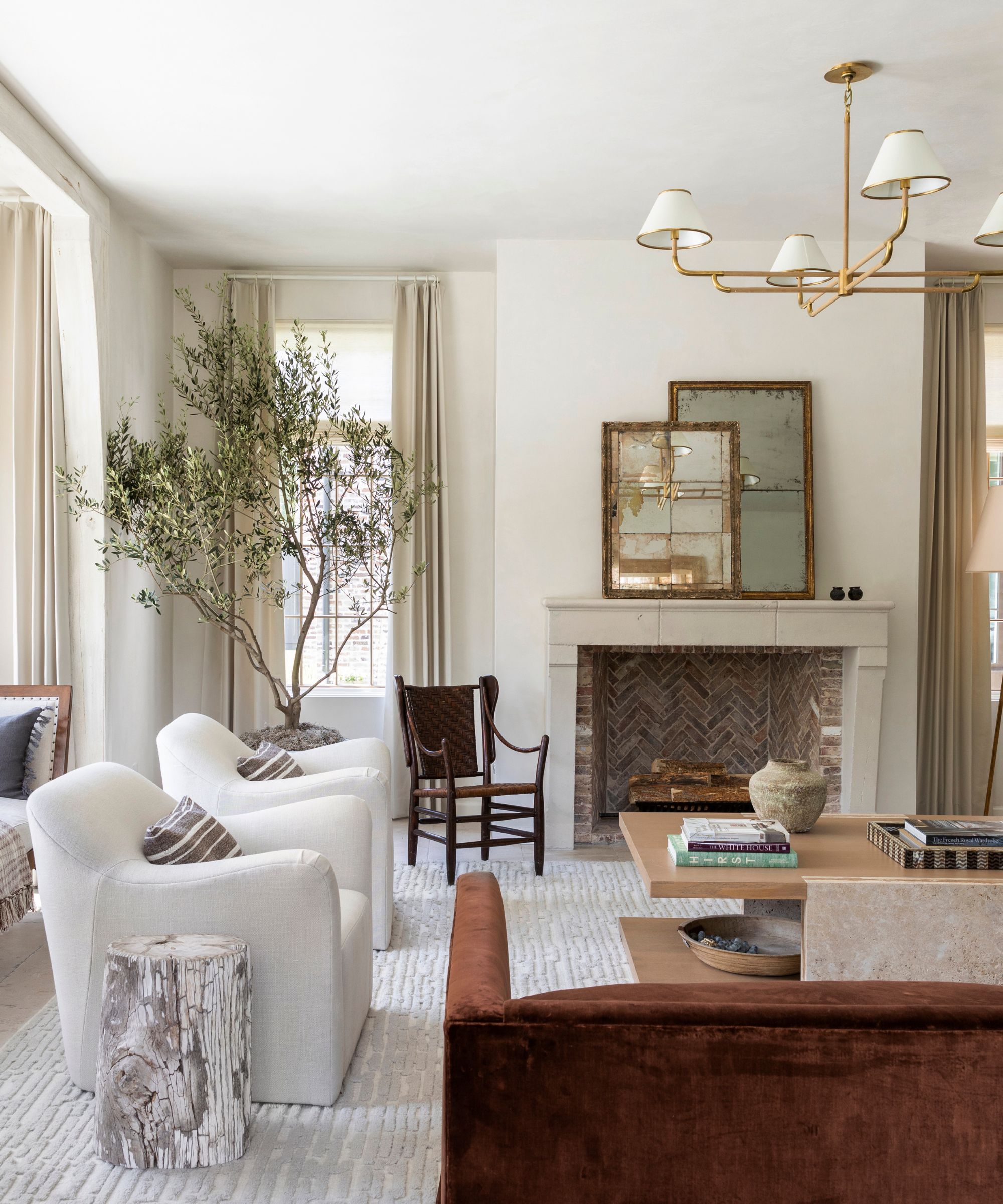 Transitional living room with soft white walls, beige curtains, a stone fireplace with antique mirrors, a light gray rug with two white armchairs, a wooden coffee table, and a rust-colored velvet sofa