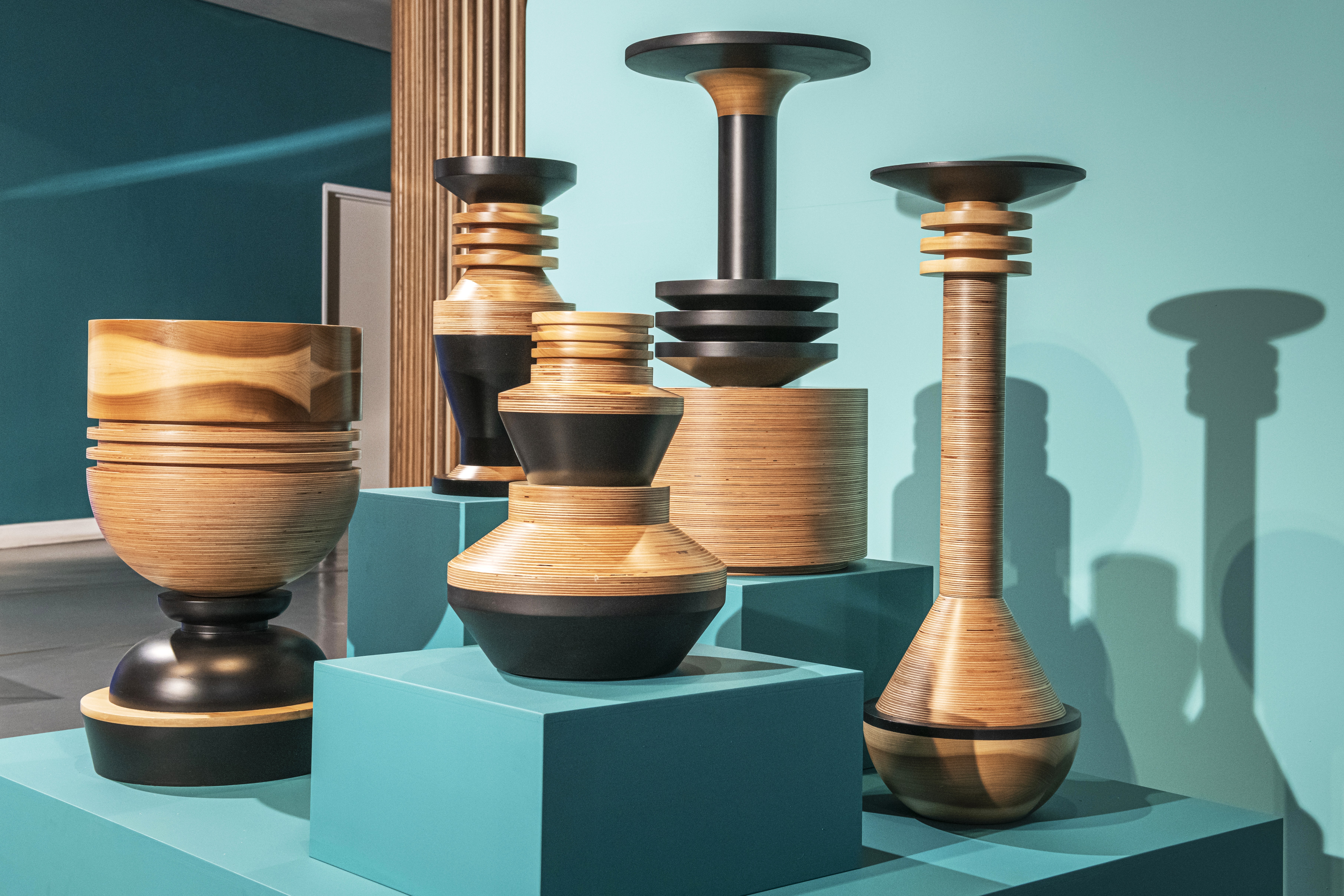 A series of highly textural wooden sculptures with a totemic structure and a dark and pale wood silhouette sit atop teal-painted podiums in a brightly lit gallery.