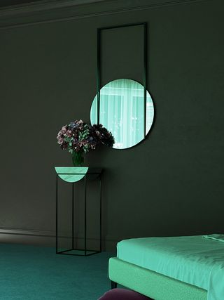 A 3D render of a room. A circular mirror is hung on the wall, below which is a vase filled with flowers, set on a metal and marble table. We see a corner of the bed.