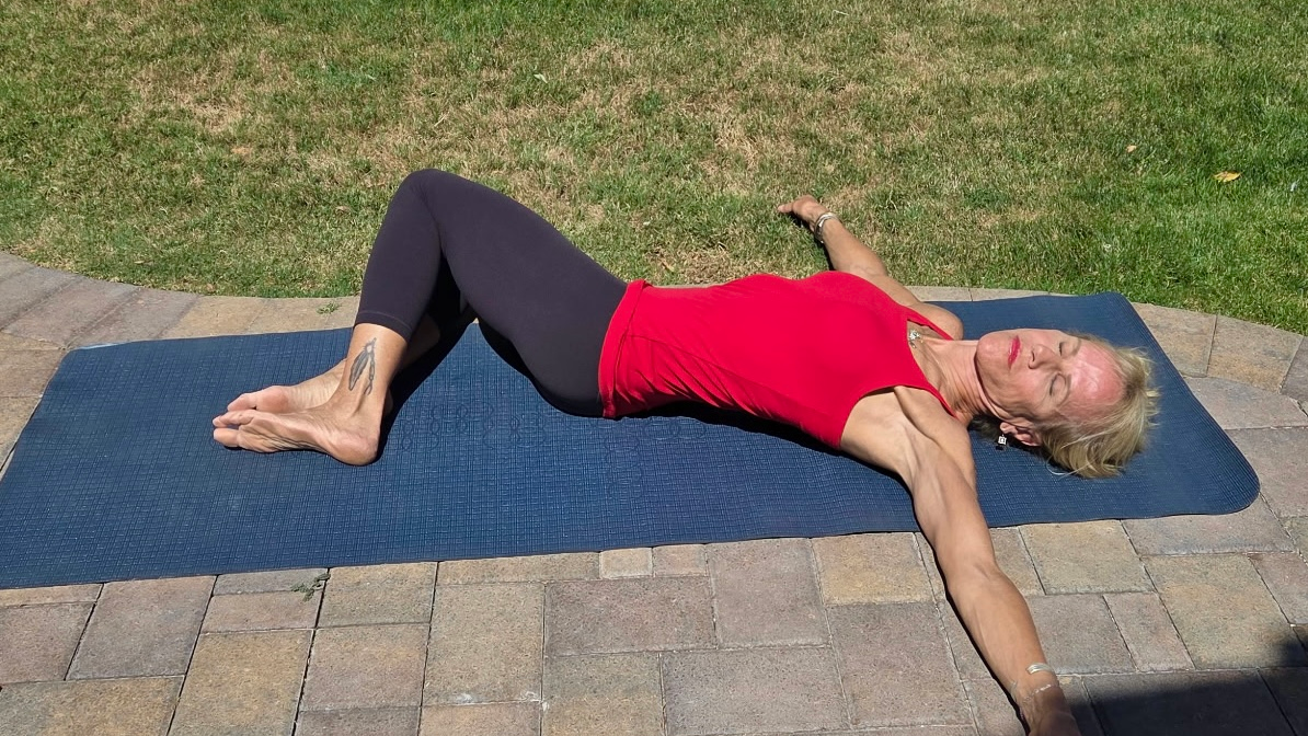 Woman in black leggings and red tank top on exercise mat on patio outside, next to grass.