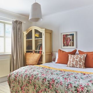 bedroom with white walls and flowered quilt and orange cushions with cream coloured wardrobe with wire mesh front