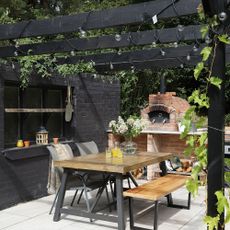 Black painted wooden pergola with string lights above outdoor dining area with wooden bench and brick kiln
