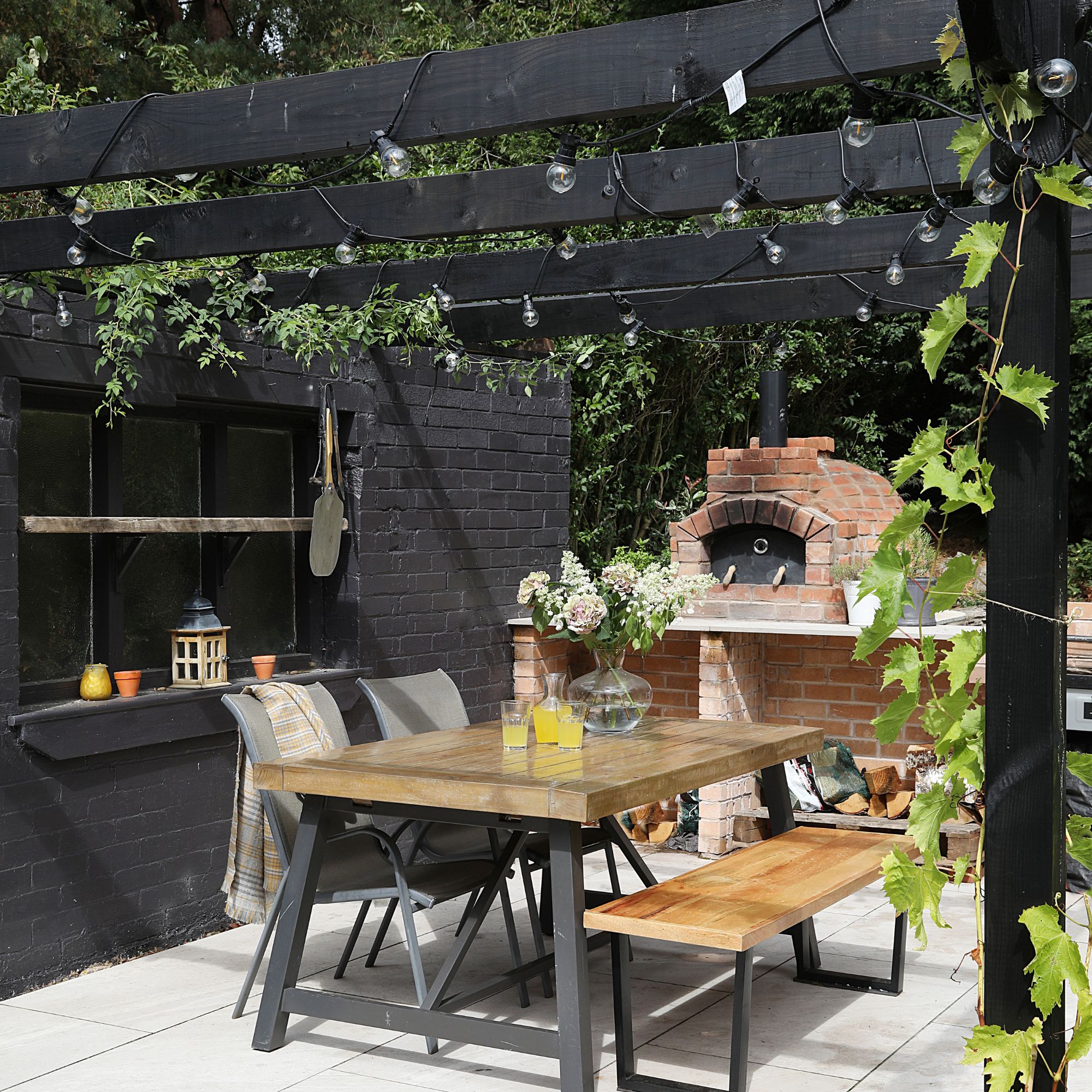 Black painted wooden pergola with string lights above outdoor dining area with wooden bench and brick kiln