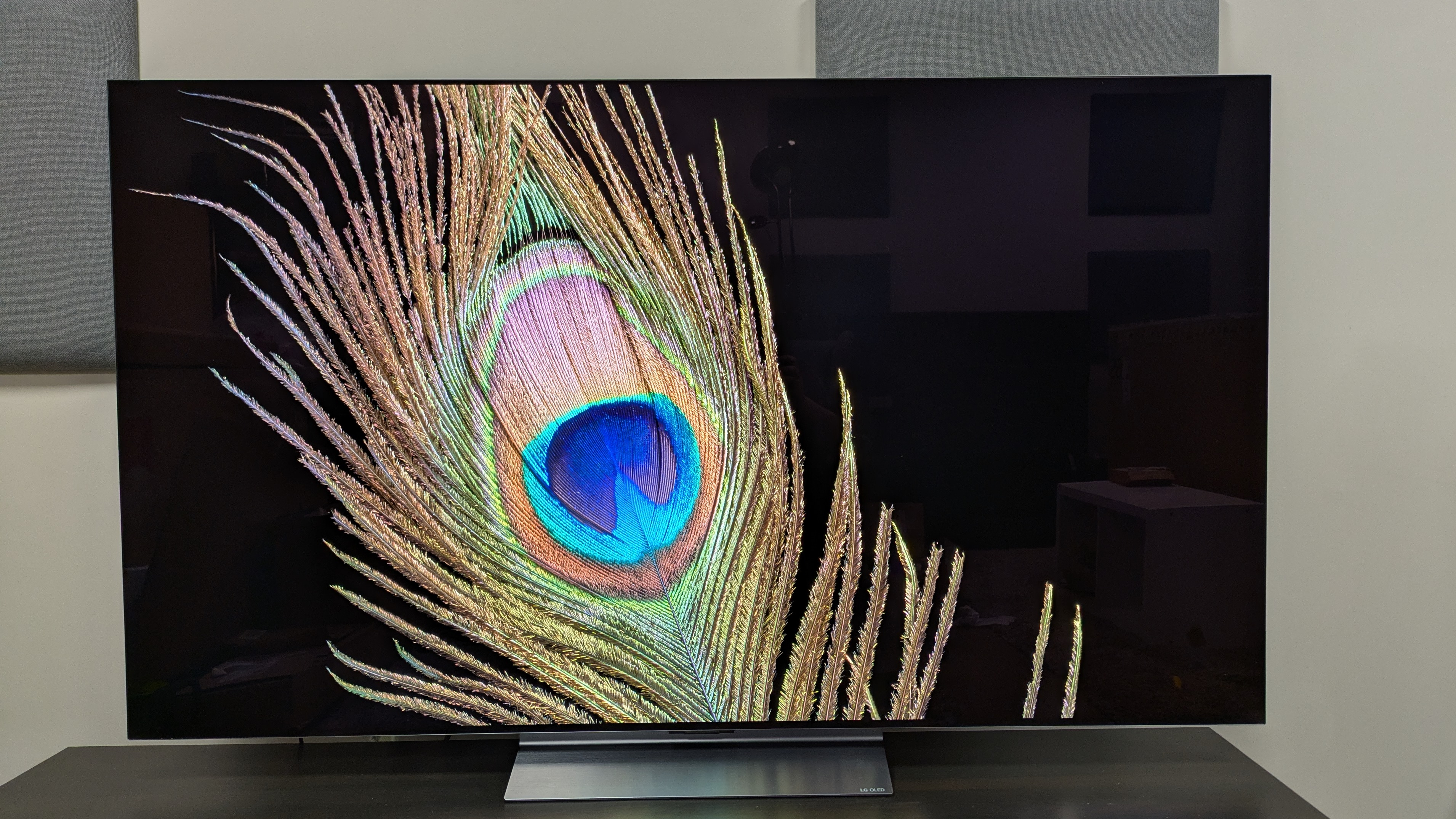 The LG C6 OLED TV with a peacock feather against a black background on screen. The peacock feather is detailed and the background is a deep black, but some reflections from the room are visible.