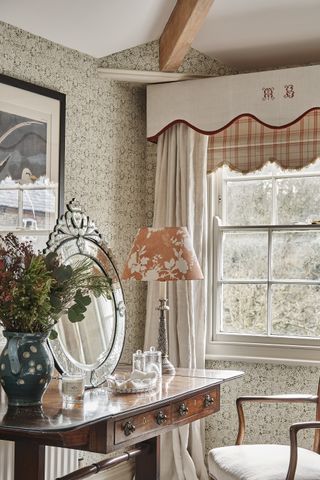 corner of a bedroom with drapes and pelmet, green wallpaper, dressing table with mirror, table lamp, artwork, vase, chair