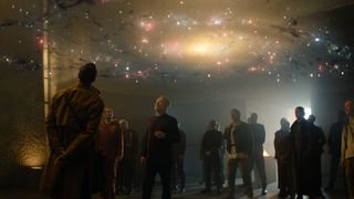 a group of people in dark clothing stand in a dimly-lit room looking up at a holographic floating model of a galaxy