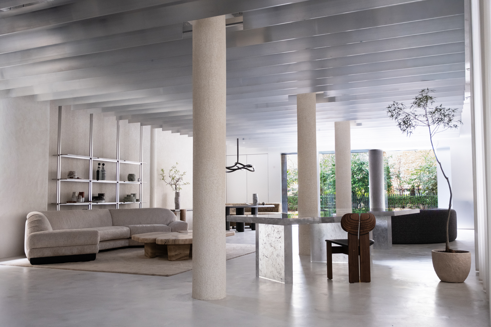 The interior of a brutalist, minial design showroom, decorated with contrasting furniture, artworks, and accessories in wood, earthy fabrics, reflective surfaces, and plants.