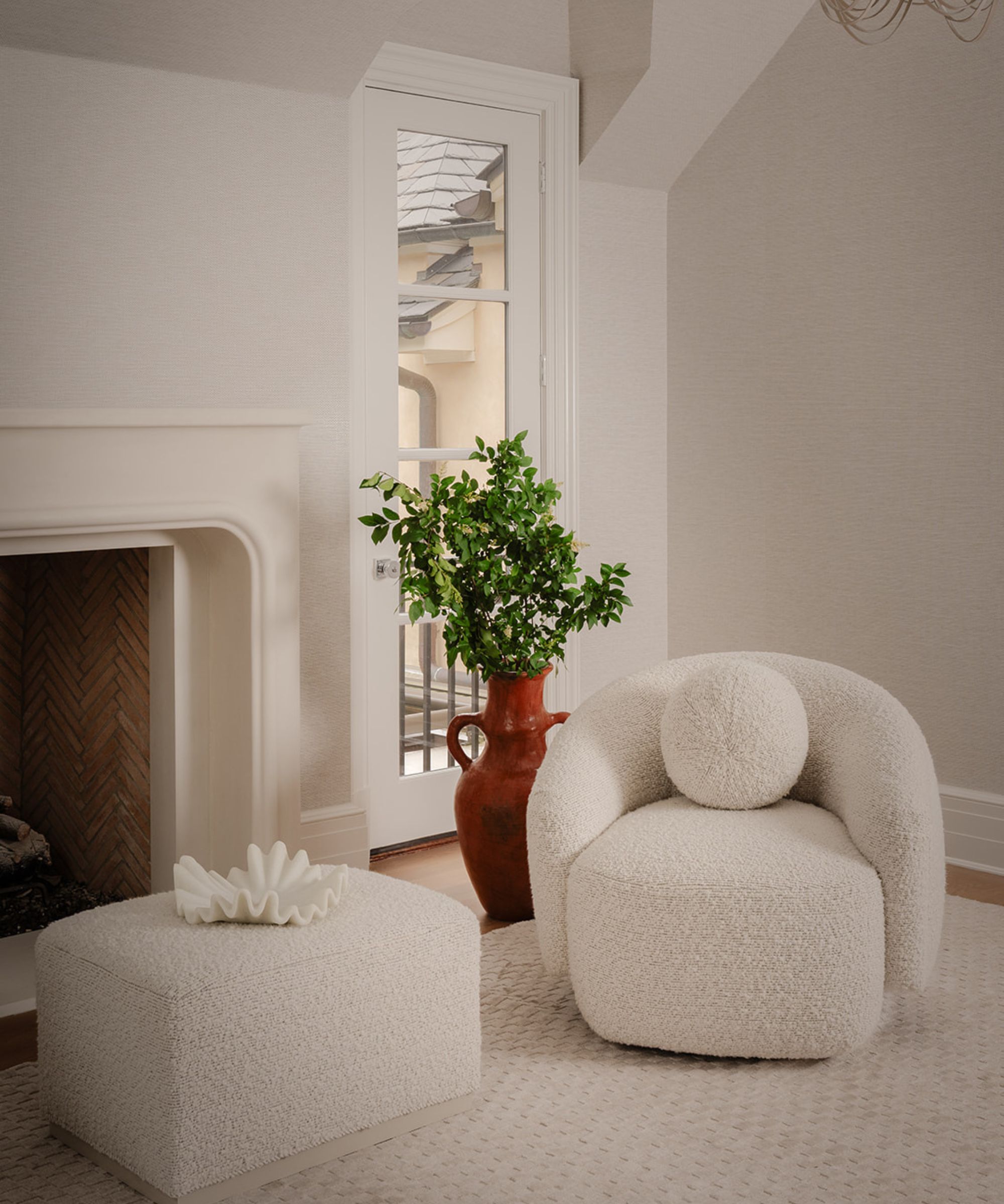Cream living room with a fireplace with a brick interior and a boucle armchair and matching pouffe and a large vase filled with greenery