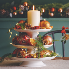  Cake stand with golden baubles, pine cones and Christmas decorations with a lit candle on the top.