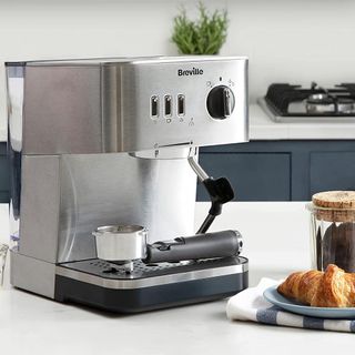 Brevill Espresso Coffee Machine on a white counter-top next to a croissant and a grounded coffee-filled jar