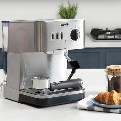 Brevill Espresso Coffee Machine on a white counter-top next to a croissant and a coffee-filled jar