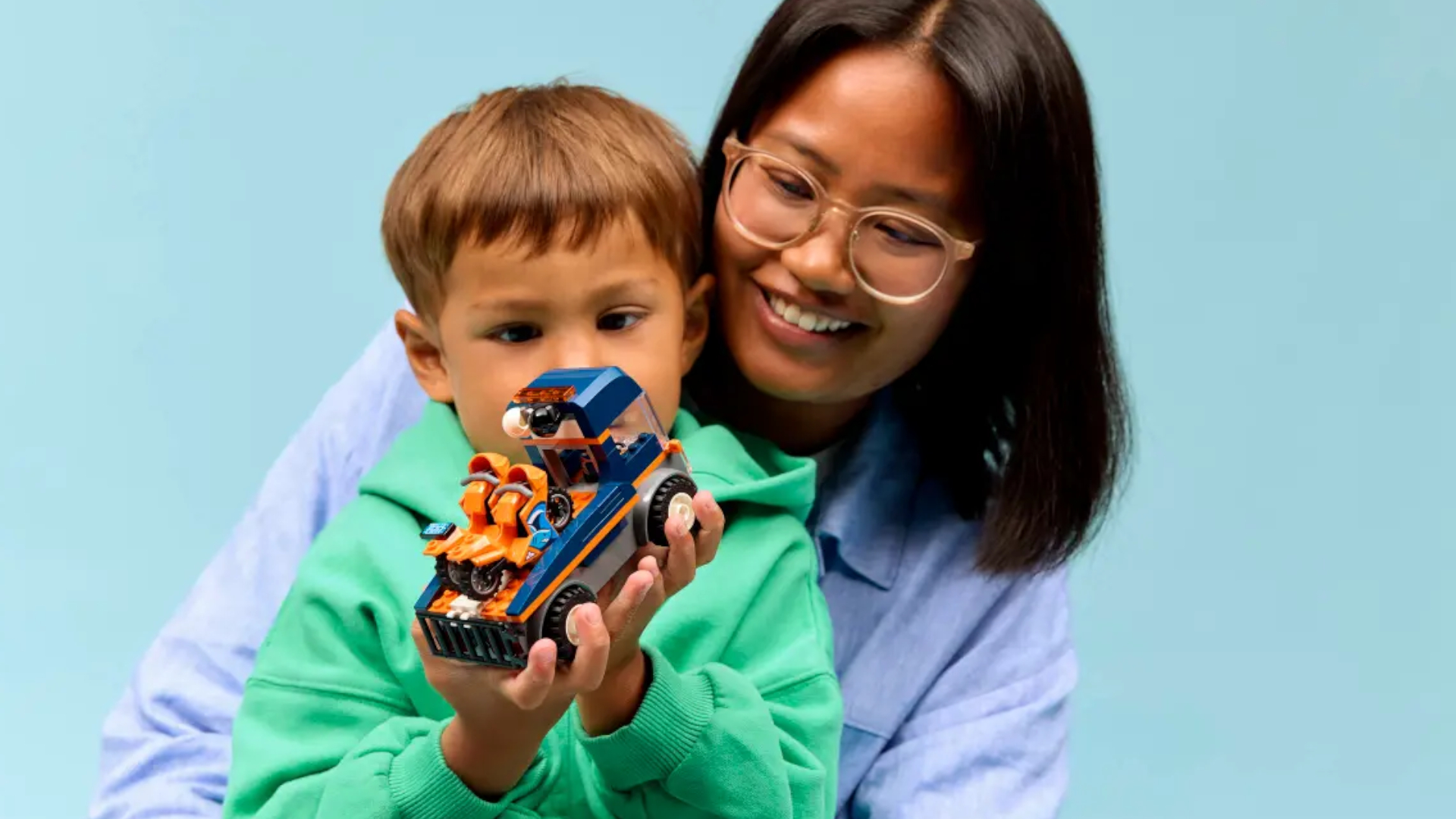 A woman holds a child who is playing with the Lego Motorcycle Transporter
