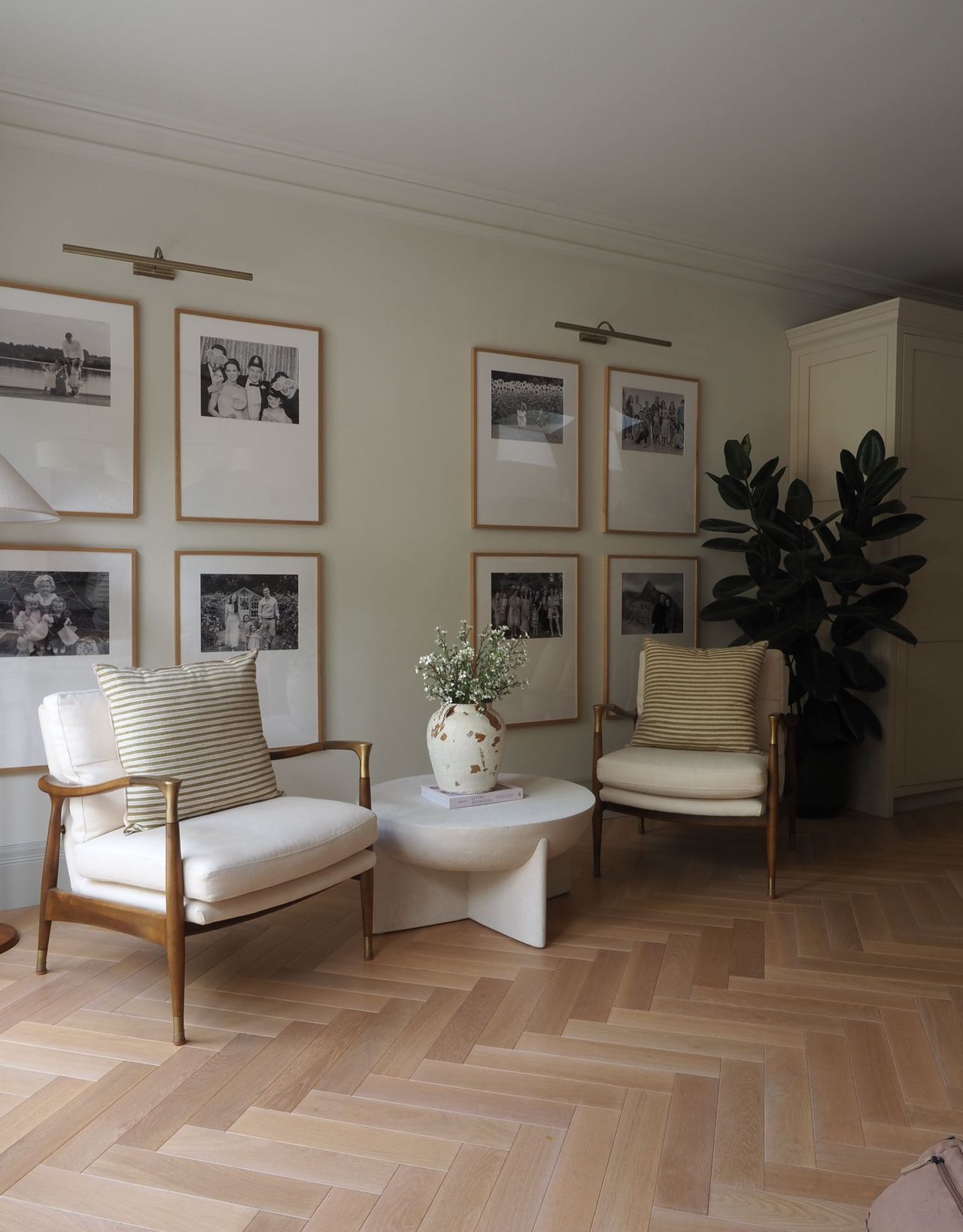 A seating area with two white-cushioned armchairs with wood legs and brass corner arm details in front of framed black and white photos with a large plant in the background