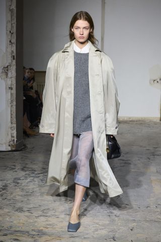 A model at Carven's spring/summer 2024 runway show wearing a trench coat with a gray sweater, a gray sheer skirt, and gray square-toe mules.