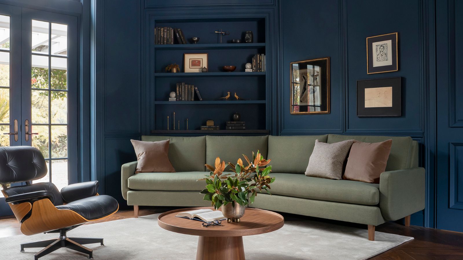 Home office with dark blue walls, curved green sofa, eames chair, wood round table and bookshelves