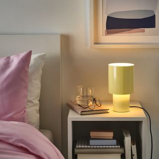 Close-up image of a bed and a bedside table in a white bedroom. There is light pink bedding on the bed and the table has a few books on it, glasses, a glass of water, and a light yellow lamp that is on.