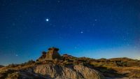 A glowing bright blue night sky full of white stars looks over a rocky landscape. 