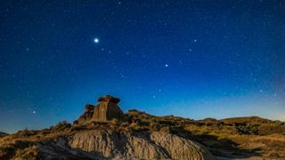 A glowing bright blue night sky full of white stars looks over a rocky landscape. 