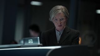 Close up of a woman with short gray hair and glasses wearing a smart suit. She is looking down intently at a screen in front of her.