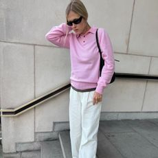 Woman wears a pink polo sweater, white baggy trousers, boat shoes and wraparound sunglasses.