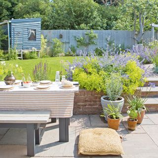 country cottage garden with outdoor dining table and benches with tablecloth, floor cushion, planters and a wooden playhouse in the garden