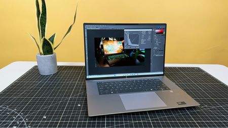 HP ZBook Studio 16 G11 mobile workstation on a white desk with a black table mat, against a yellow backdrop.