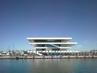America’s Cup Building is set next to a body of water. This modern building has four protruding white constructions that separate the floors. Each floor has panoramic windows.