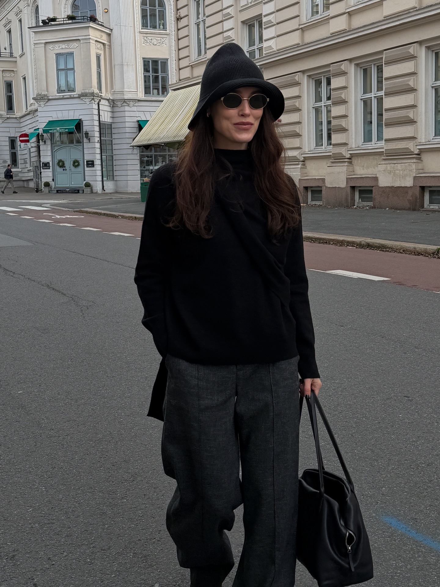 An image of @annabelrosendahl wearing a black hat from The Row, a scarf jumper, grey pleated trousers and an oversized bag while out in Norway.