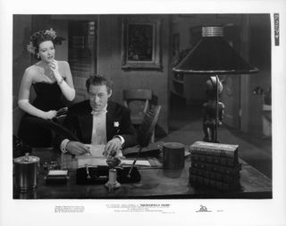 a woman in evening wear stands by a man in a suit at his desk in the movie unfaithfully Yours