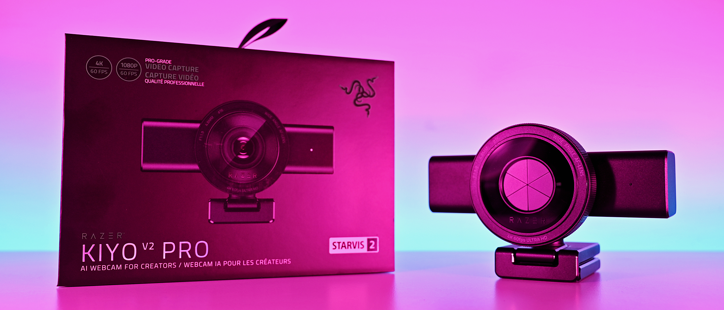 Wide shot of the Razer Kiyo V2 Pro webcam with lens shutter closed, next to its box under pink and blue ambient light.