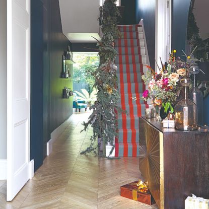 A navy-painted hallway with a staircase covered in a red and grey striped runner and the bannister covered with a garland