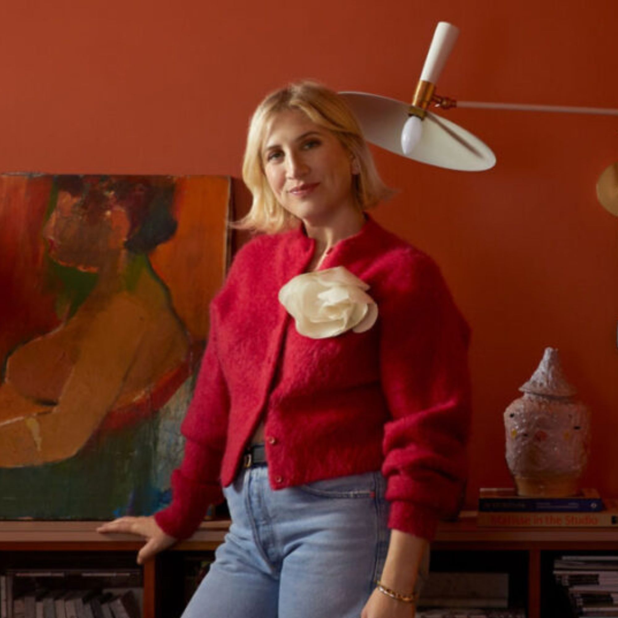 woman with short blonde hair and red cardigan in a room painted deep orange with an artwork and sculpture behind her and a wall lamp